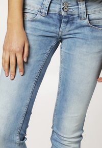 Jeans de mezclilla azul claro con un ajuste Slim, que presentan un lavado desgastado, cinco bolsillos y dos botones metálicos en la cintura.