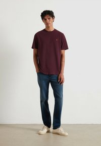 T-shirt in cotone bordeaux con collo rotondo e maniche corte, abbinato a jeans in denim blu e sneakers bianche. Dettaglio del logo ricamato in modo sottile.