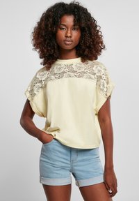 Gul T-shirt med blommig spetsdetalj på axlarna, kortärmad, avslappnad passform. I kombination med ljusblå denimshorts med uppvikta fållar.