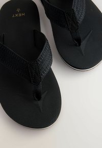 Mustat flip-flopit, joissa on tekstuuripinnalla varustetut hihnat ja sileä pohja. Hihnoissa on kudottu kuvio ja pieni musta korostus keskellä.
