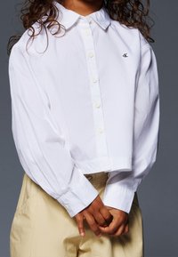 Witte geknipte knopshirt met een kraag, lange mouwen en een geborduurd logo. Gepaarde met lichtbeige broek, die een casual look laat zien.