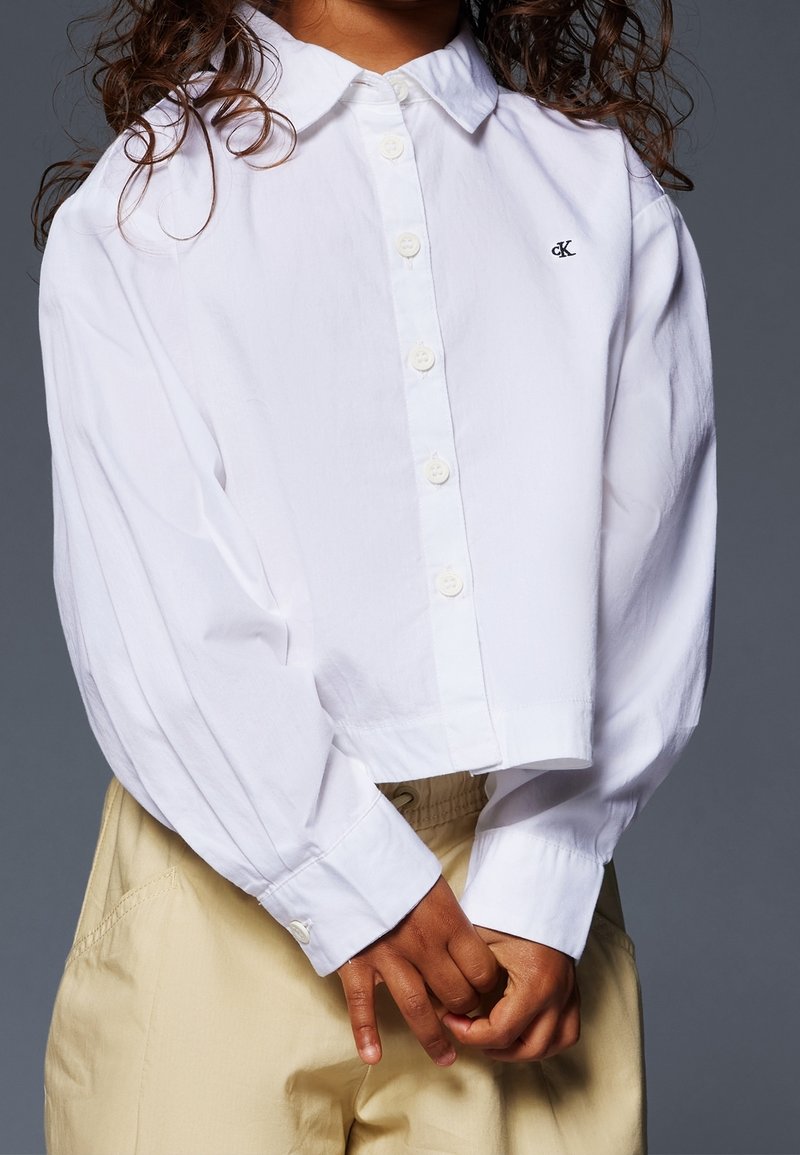 Witte geknipte knopshirt met een kraag, lange mouwen en een geborduurd logo. Gepaarde met lichtbeige broek, die een casual look laat zien.