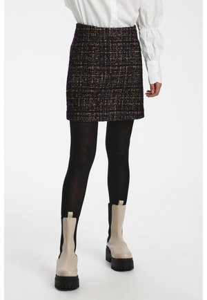Mini-jupe en tweed noir avec une surface texturée, présentant une taille haute. Portée avec des collants noirs et des bottines beiges.