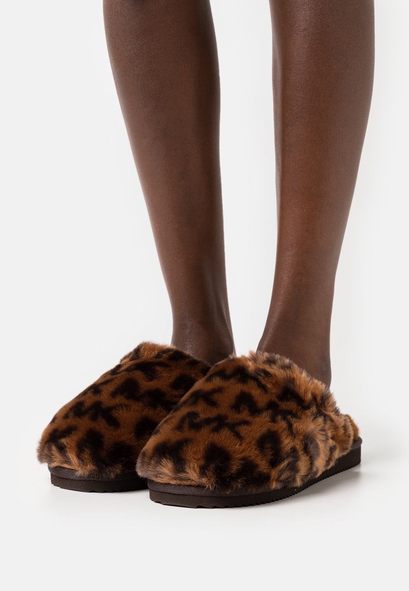 Chaussons bruns en peluche avec motif léopard, texture douce, design à bout ouvert et semelle en caoutchouc noire, posés sur des jambes nues sur fond blanc.