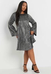 Silverfärgad metallisk klänning med vertikala plisseringar, långa utsvängda ärmar, rund halsringning och en svag kurva vid fållen. Kombinerad med svarta remsandaler.