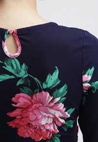 Mörkblå klädesplagg med stort rosa blommönster. Bakre stängning med en liten nyckelhålsgrop och knapp. Mjuk tygstruktur som är tydlig.