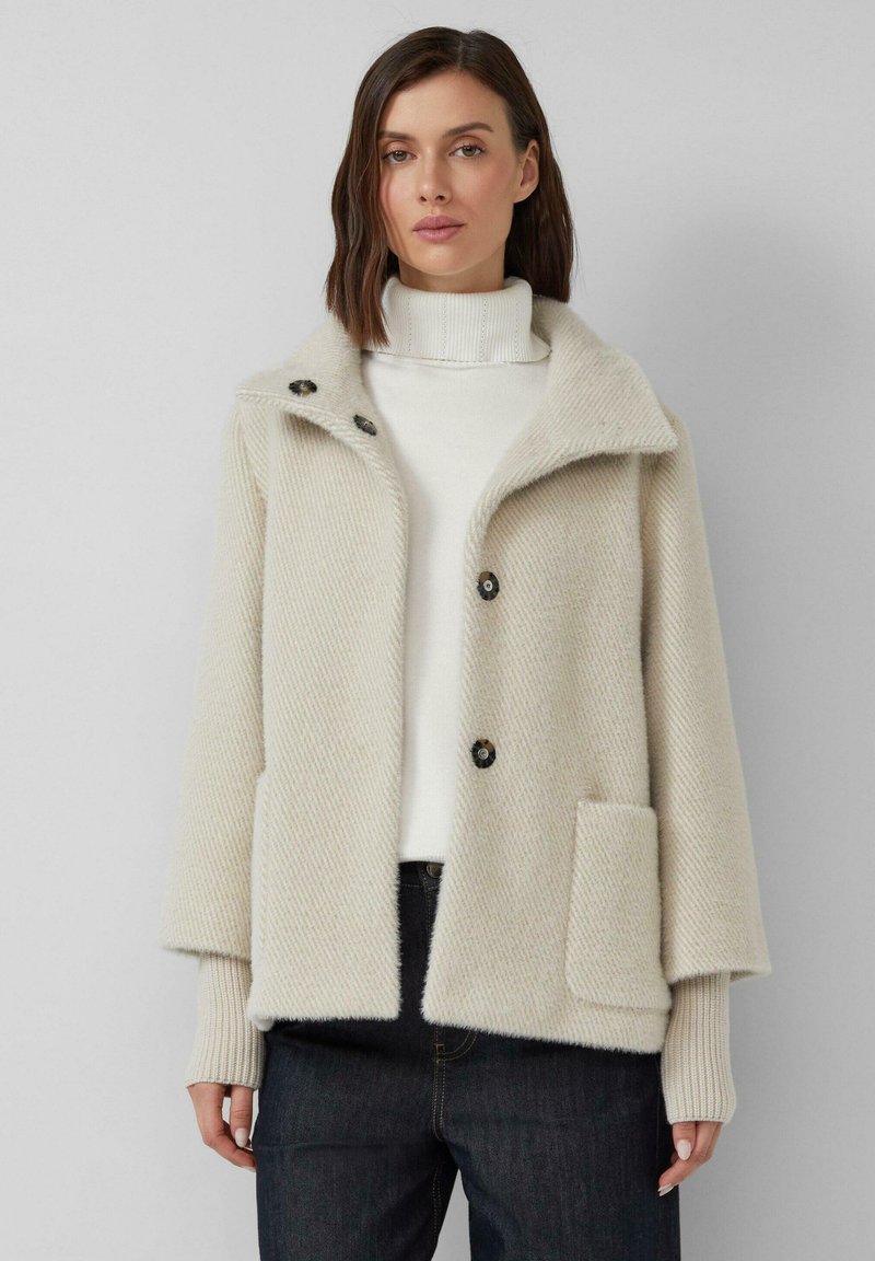 Licht beige, gestructureerde wollen mix jas met een brede kraag, drie zwarte knopen en een voorzak. Draagt over een witte turtleneck.