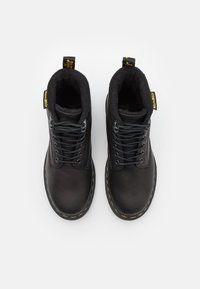 Botas de cuero negro con un acabado suave, puntera redonda, cordones grises, forro negro suave y costuras amarillas alrededor de la suela.