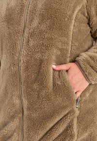 Fleece takki vaaleanruskeana, jossa on vetoketjukiinnitys, teksturoitu pinta, sivutaskut ja rento istuvuus. Pehmeä materiaali, jossa on ylellinen tuntu.