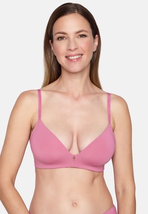 Lächelnde Frau mit glatten braunen Haaren, die einen pinken BH mit dünnen Trägern vor einem einfarbigen hellen Hintergrund trägt.