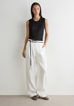 Femme portant un haut noir sans manches, un pantalon blanc à jambes larges avec une ceinture noire, et des chaussures marron à talons, debout contre un mur clair uni.