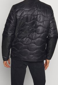 Giacca a vento nera con motivo a onda trapuntato, realizzata in materiale lucido. Presenta un collo rotondo e maniche lunghe.