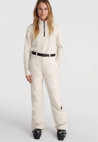 Vrouw staat in een crèmekleurig zip-nektrui, bijpassende broek met een zwarte riem en zwarte skibotten op een effen lichte achtergrond.