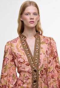 Robe florale en tissu rose pâle avec des finitions vertes, mettant en avant des manches bouffantes, un devant boutonné et des motifs complexes de fleurs et d'animaux.