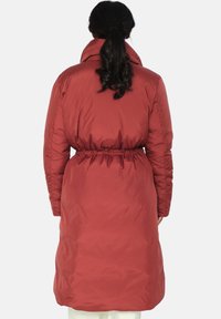 Rød polstret kåpe med høy krage, innsvingt midje og mansjetter ved håndledd. Jevn tekstur med blank finish, knelengde design.