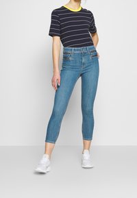 Žena nosící světle modré džíny s vysokým pasem a předními zipy, námořnicky pruhované tričko se žlutým límečkem a bílé tenisky.