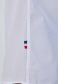 Tissu blanc avec une couture verticale et une petite étiquette en tissu présentant des rayures horizontales bleu marine, blanches et rouges près de la couture.