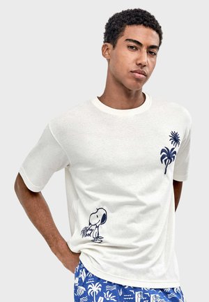 Jonge man draagt een wit T-shirt met marineblauwe palmboom- en Snoopy-ontwerpen, gecombineerd met blauwe, met patronen bedrukte shorts, staand tegen een effen achtergrond.