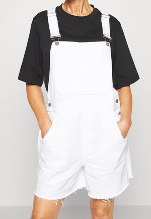 Witte spijkerbroek-overalls met korte pijpen, voorzien van grote voorzakken en verstelbare banden, gedragen over een zwart shirt met korte mouwen.