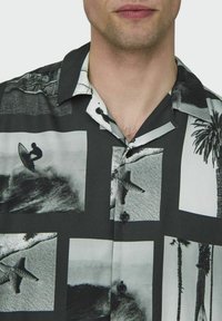 Camisa de manga corta en tejido ligero con estampados fotográficos en blanco y negro de surf y palmeras, que cuenta con cuello y botones.