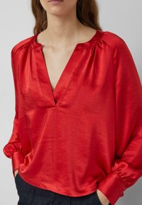 Frau trägt eine rote Satin-V-Ausschnitt-Bluse mit langen Puffärmeln und breiten Manschetten, kombiniert mit dunkler Hose, Hände in den Taschen.