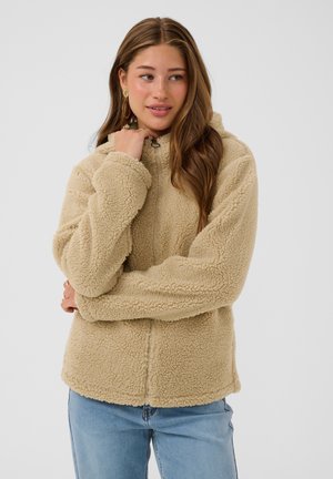Young woman with long brown hair wearing beige fluffy zip-up jacket and blue jeans against a plain light background.