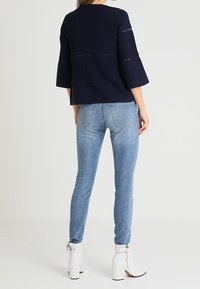 Tummansininen ribattu neulontoppi, jossa on kellotetut hihat, yhdistettynä vaaleansinisiin skinny-farkkuihin ja valkoisiin nilkkureihin, joissa on vetoketjut ja hopeinen varuste.
