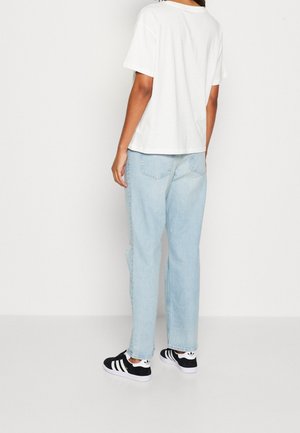 Persoon die een effen witte korte mouw T-shirt, lichtblauwe high-waisted jeans en zwart-witte Adidas sneakers draagt, staand met de rug naar voren.