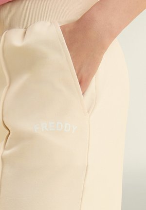 Main reposant dans la poche avant d'un pantalon beige avec une ceinture côtelée et le logo "FREDDY" sur la cuisse.