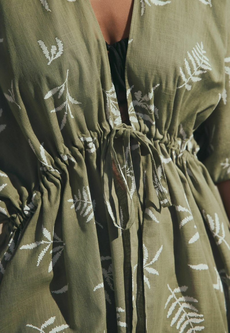Gros plan d'une robe verte avec des motifs de feuilles blanches, présentant une taille froncée nouée avec un nœud en tissu sur un vêtement intérieur noir.