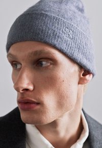 Berretto lavorato a maglia grigio realizzato in materiale morbido, con una texture a coste e un piccolo dettaglio del logo sul lato. Vista ravvicinata di un modello maschile.