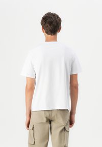 T-shirt en coton blanc à manches courtes, col rond, texture lisse, portée avec un pantalon cargo beige comportant plusieurs poches.