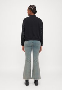 Veste bomber noire avec des manches en sequins, associée à un jean bleu clair évasé et des bottines noires à talons. Fond uni.