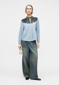 Kvinde iført lyseblå stribet bluse med sort blonder yoke, vide jeans, sorte sko, og holder lille beige håndtaske.