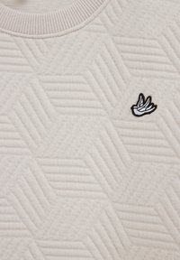 Krém színű pulóver texturált geometriai mintával. Kerek nyakkal és egy kis, fekete-fehér hímzett madár logóval.