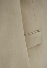 Zbliżenie na beżowy materiał, ukazujące gładką fakturę i subtelny połysk, z eleganckim detalem kieszeni i precyzyjnym stębnowaniem wzdłuż krawędzi.