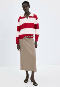 Donna che indossa una polo a maniche lunghe a righe rosse e bianche, gonna midi beige, sandali con tacco neri e una cintura decorativa.