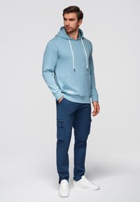 Felpa azzurra con tasca frontale, coulisse e orlo a costine, abbinata a pantaloni cargo blu navy con tasca laterale con zip. Sneakers bianche.