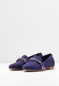 Des ballerines en daim violet présentent une lanière tressée avec des accents floraux sur le dessus. Les chaussures ont un bout rond et une semelle brune basse.
