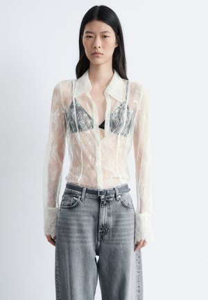 Translucent lace shirt with a detailed floral pattern, button-down front, pointed collar, and long sleeves. Worn over a black bralette and gray jeans.