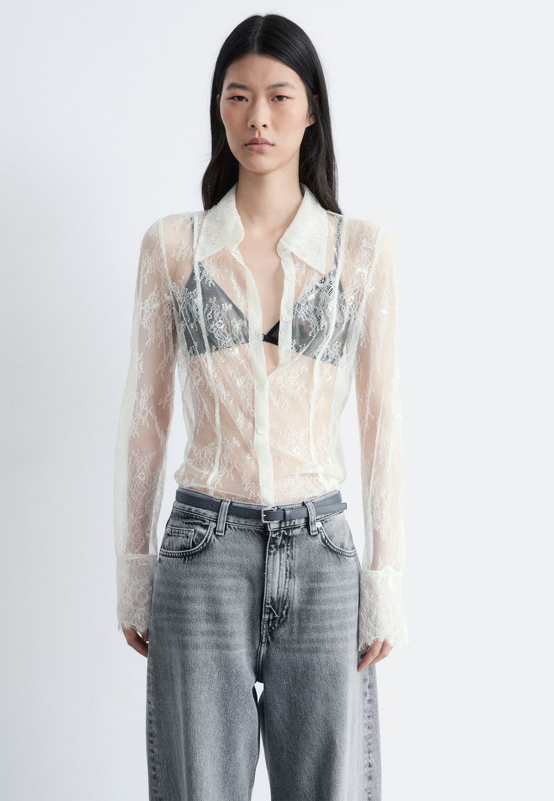 Chemise en dentelle translucide avec un motif floral détaillé, fermeture boutonnée, col pointu et manches longues. Portée sur un bralette noire et un jean gris.