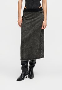 Γυναίκα με μαύρο αμάνικο τοπ, γκρι μίντι φούστα με πλευρικές ρίγες και μαύρα μπότες με τακούνι και μυτερό μύτη, σε λευκό φόντο.