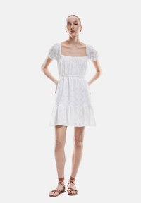 Vestido de algodón blanco con mangas abullonadas, escote cuadrado y dobladillo con volantes. Patrón de ojales en toda la prenda que añade textura. Combinado con sandalias marrones.