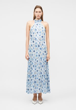 Kvinde iført en ærmeløs, højhalset, ankel-lang kjole med blå blomsterprint og hvide spidse hæle mod en ensfarvet baggrund.