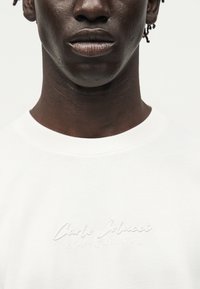 Gros plan d'une personne portant une chemise blanche avec un texte blanc brodé subtil « Carlo Colucci A Legacy Since 1976 » sur la poitrine.