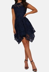 Navy blue lace dress with a fitted bodice, short sleeves, and an asymmetrical, layered hem. Features floral patterns and transparent overlay.