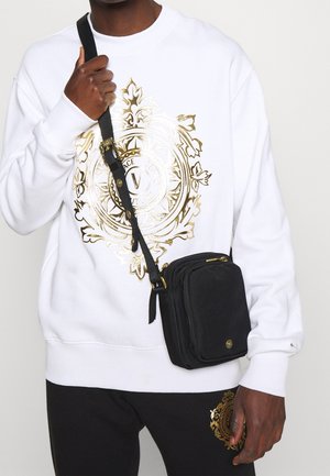 Personne portant un sweat-shirt blanc et un pantalon noir avec un emblème doré orné, tenant un sac bandoulière noir avec une fermeture éclair et des accessoires dorés.