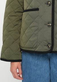 Quiltet grøn jakke med sort kant, der har store knapper og en forlommer. Ærmerne er brede, og stoffet ser glat ud.
