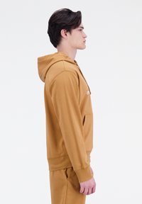 Sudadera de color mostaza hecha de tela suave, con un bolsillo canguro y puños acanalados. Modelo mostrado en vista lateral.