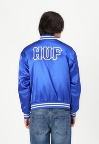 Jachetă bomber din satin albastru, cu manșete și talie din tricot cu dungi albe. Logo-ul "HUF" brodat în alb pe spate. Croi lejer.